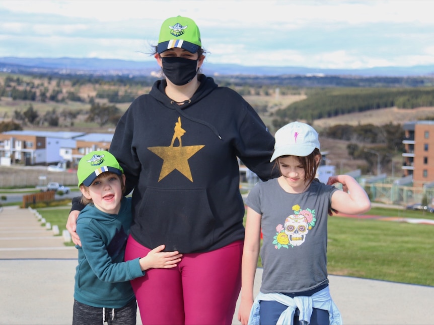 A mother with a Canberra Raiders cap wearing a mask pictured with her two children.