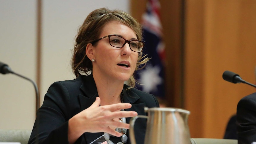A woman with dark blonde hair, glasses and a black jacket sits at a table in Senate Estimates.