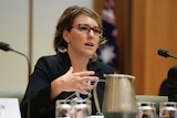 A woman with dark blonde hair, glasses and a black jacket sits at a table in Senate Estimates.