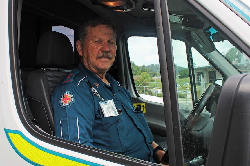 A man in dark blue overalls sits behind the wheel of an ambulance.