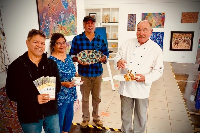 Four people posing in an art gallery with brochures, coffee, artwork and food