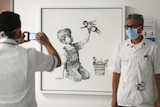 A masked health worker stands next to a painting of a young child playing with a nurse doll wearing a mask and cape