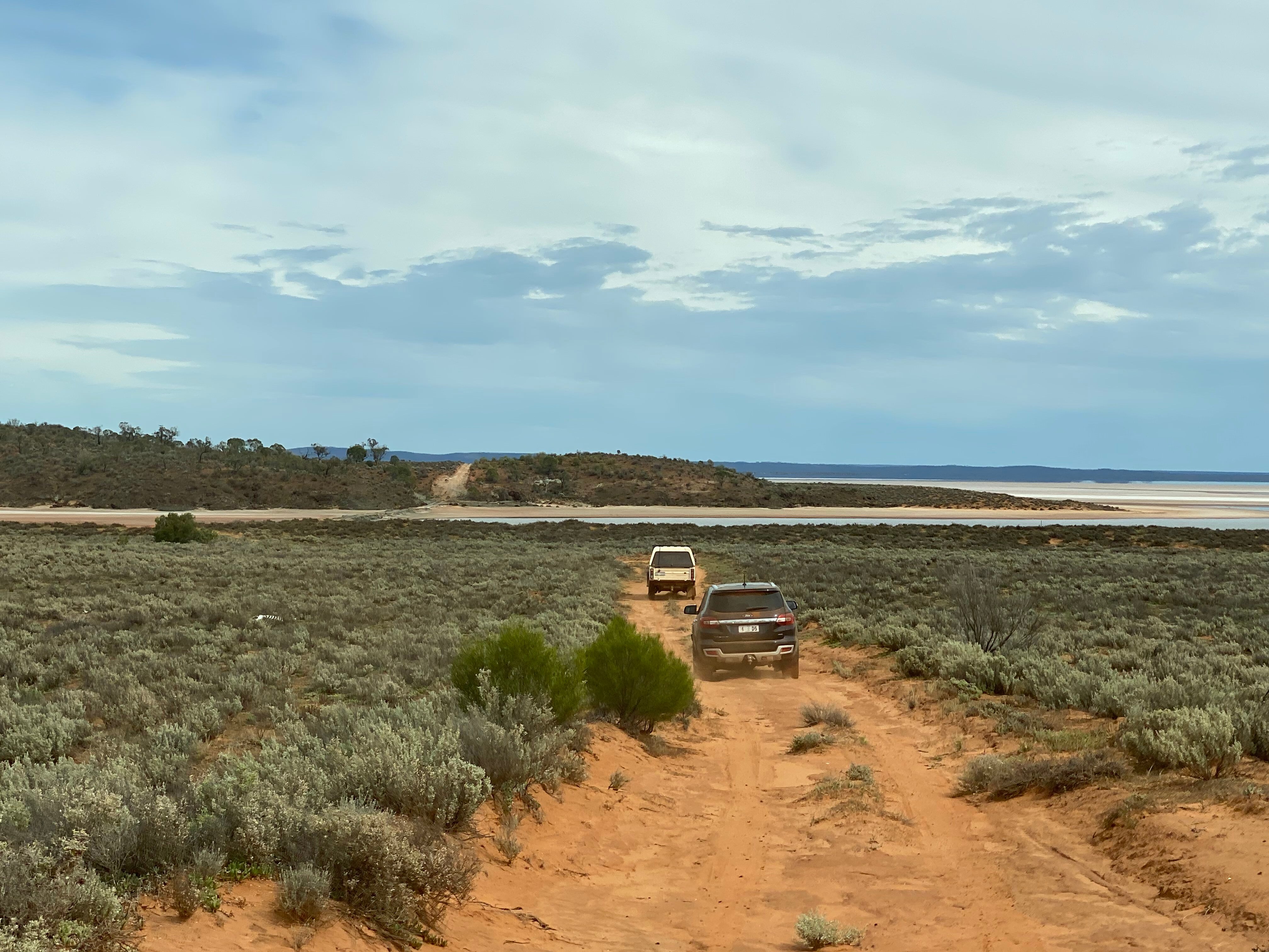 Two 4WD vehicles driving on sandy track towards a salt lake in the distance.