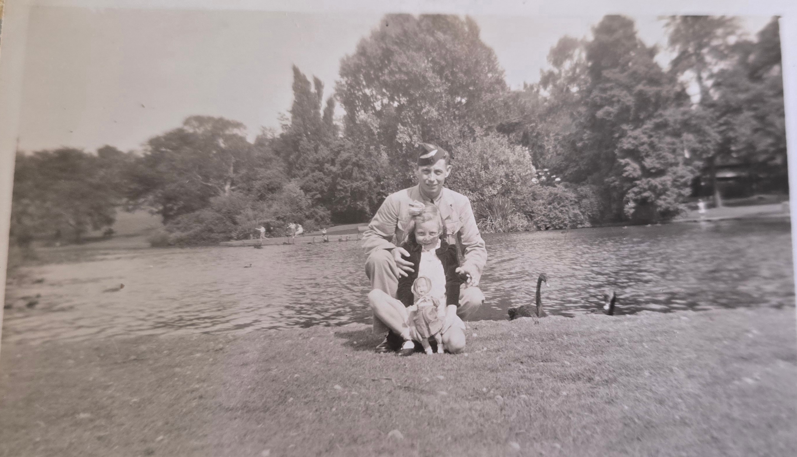 Uma fotografia sépia de um homem com uniforme da RAAF com uma menina e sua boneca. Eles estão perto da beira de um curso de água.
