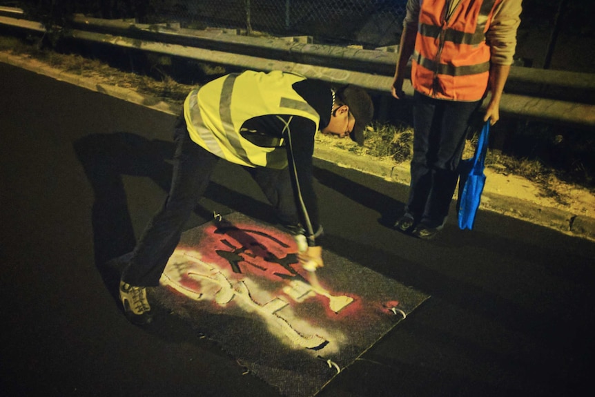 A man in a tracksuit and hi-vis vest spray paints a bike priority marking on a road using a stencil with another person nearby.