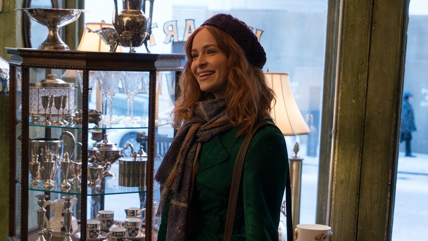 A woman wearing beret and scarf smiles and stands in the entrance of a show selling antiques and trinkets.