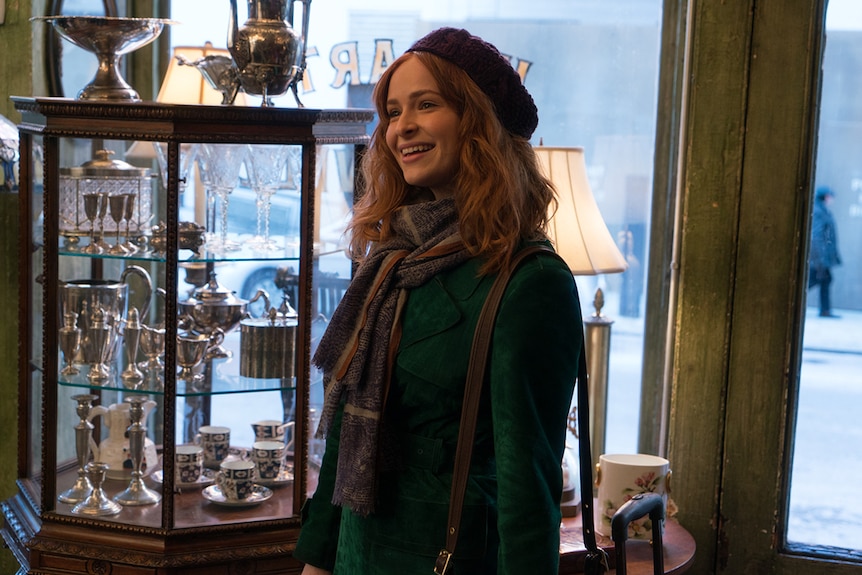 A woman wearing beret and scarf smiles and stands in the entrance of a show selling antiques and trinkets.