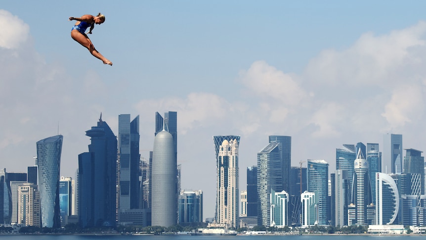 a female diver hovers in the air during a high dive with Doha skyscrapers visible behind her
