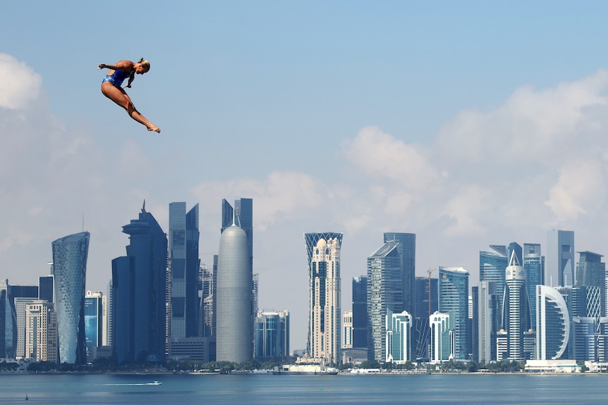 a female diver hovers in the air during a high dive with Doha skyscrapers visible behind her