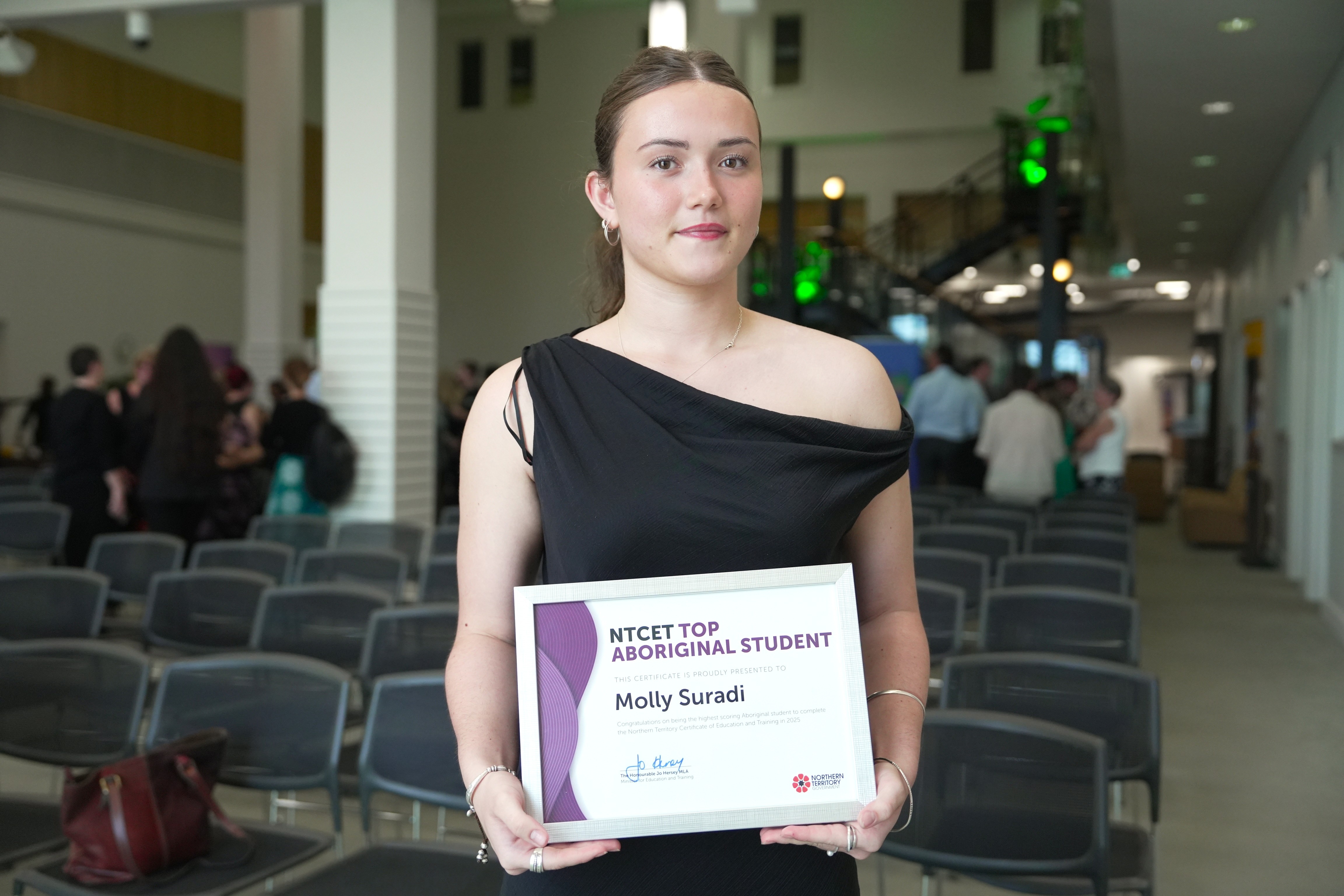 Una mujer joven, de tez clara, cabello castaño recogido en una cola de caballo, vestida con un vestido negro y sosteniendo un certificado en la mano.
