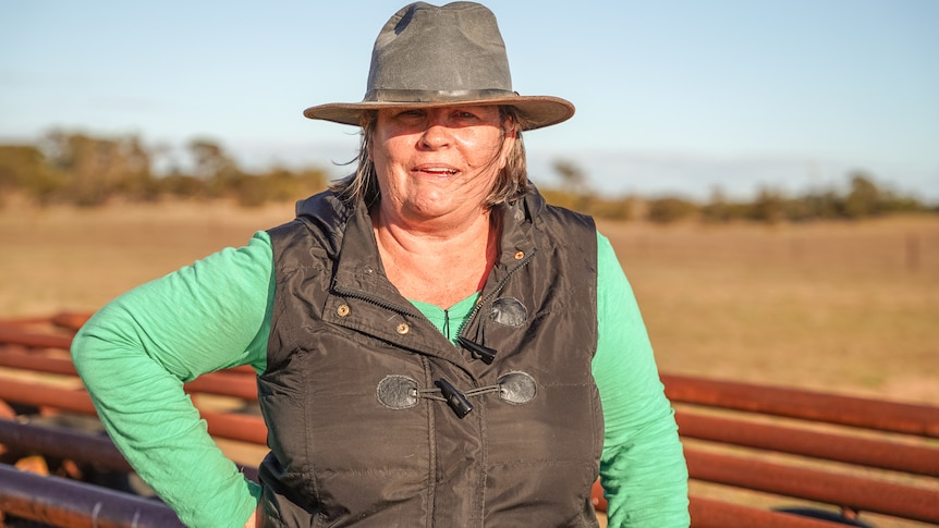 A woman wearing a long green shirt, vest and brimmed hat stands in a paddock at sunset.