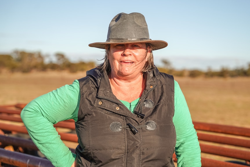 A woman wearing a long green shirt, vest and brimmed hat stands in a paddock at sunset.