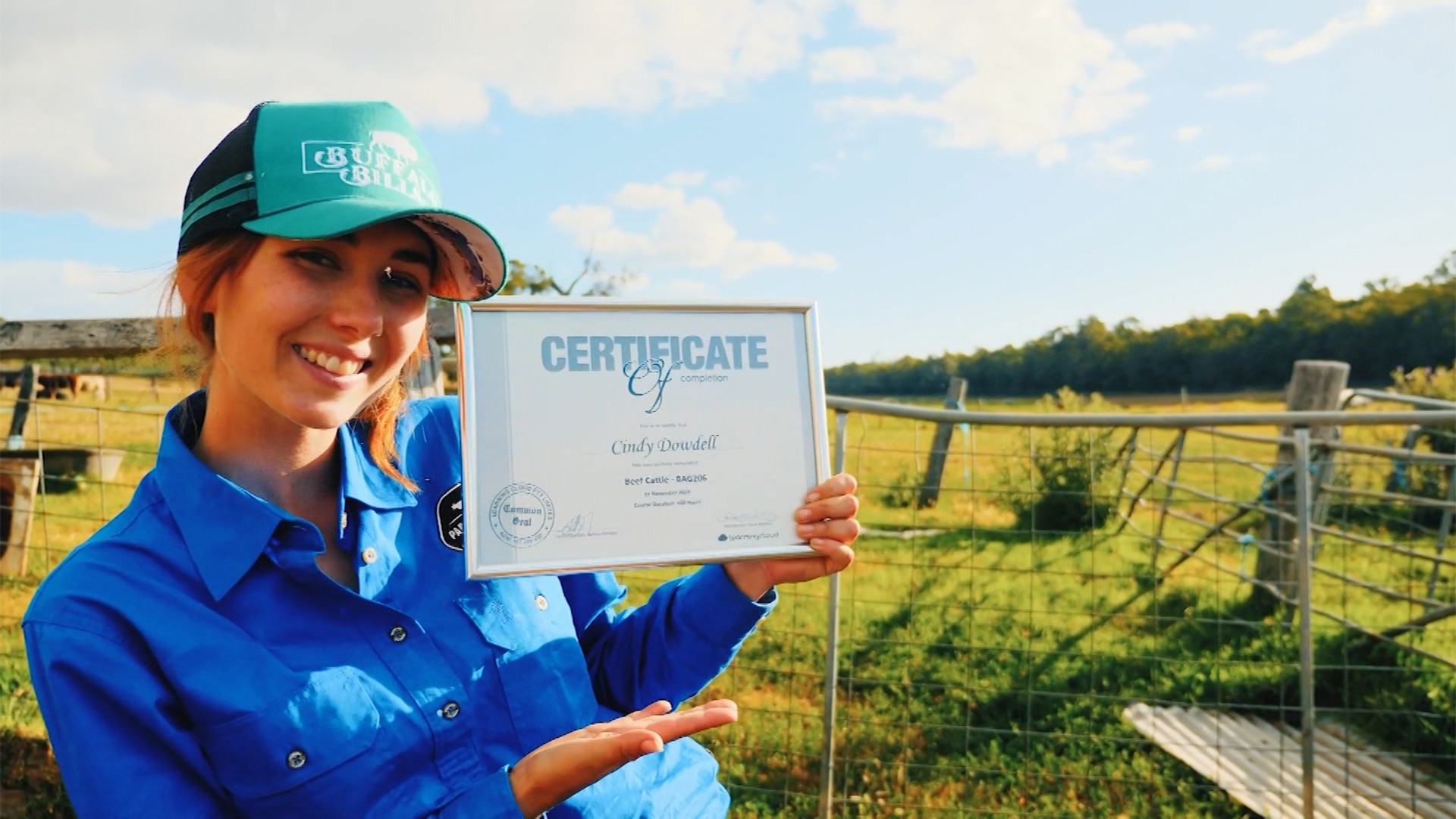 A woman stands in a field with a farm gate behind her, while wearing a blue hat and shirt and smiles as she holds a certificate