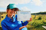 A woman stands in a field with a farm gate behind her, while wearing a blue hat and shirt and smiles as she holds a certificate
