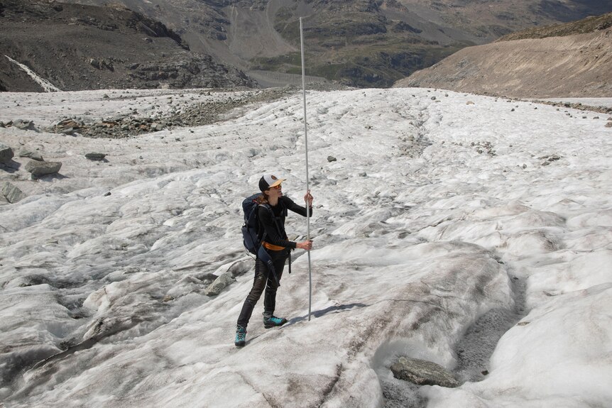 A woman holds a long rod to the ground while standing on a glacier. 