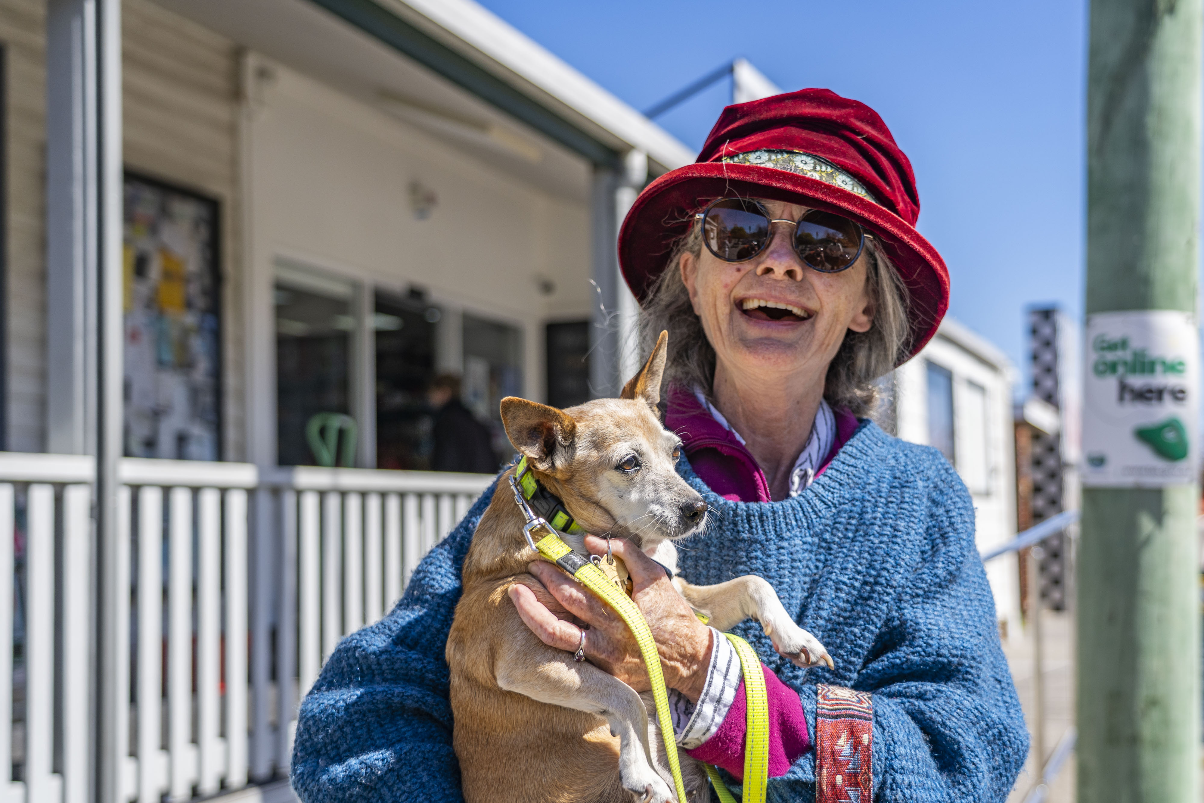 Carmel Sullivan wears a red felt hat and blue shirt and holds her small dog outside in the sun.