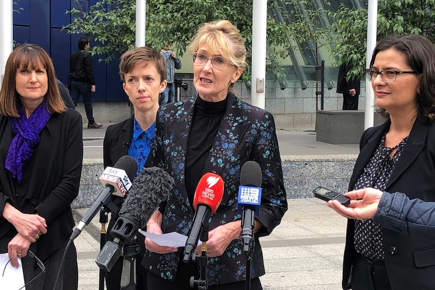 A blonde woman wearing glasses talks to the media.