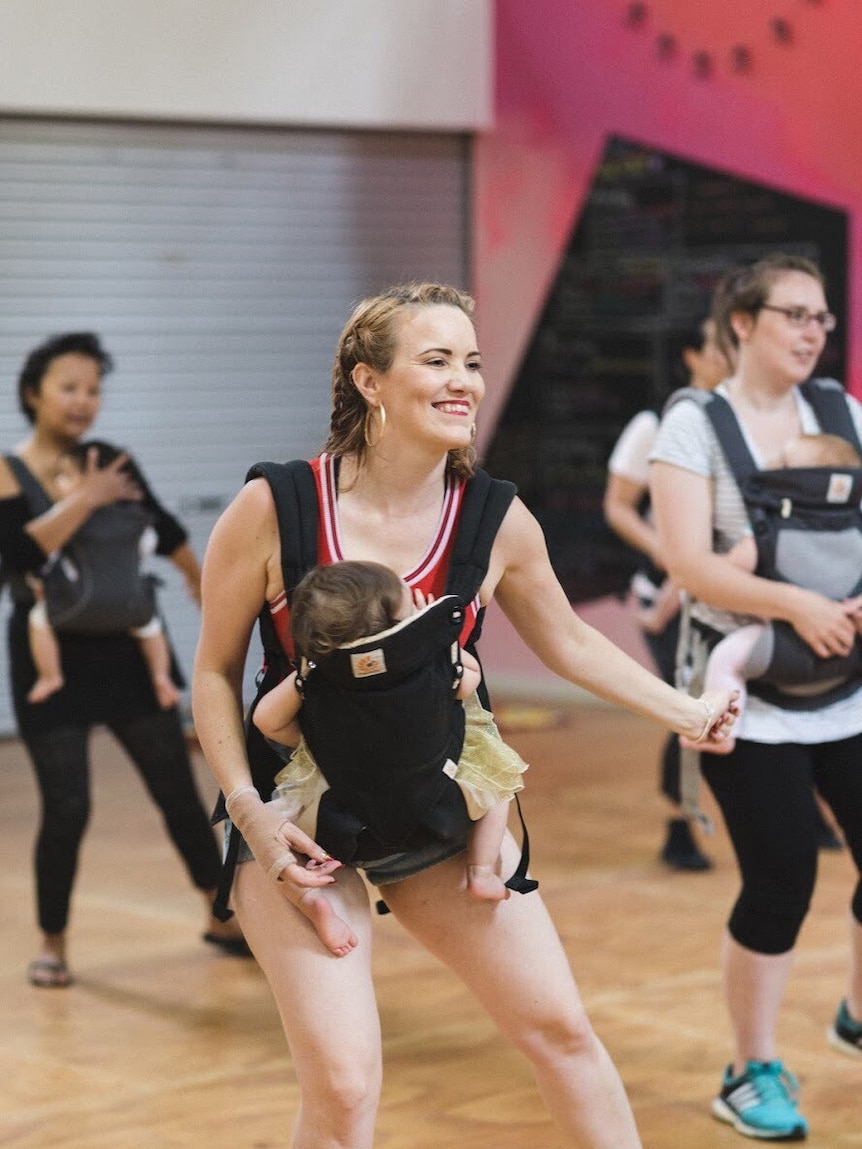 Woman leads a mothers' dance class with a baby strapped to her chest