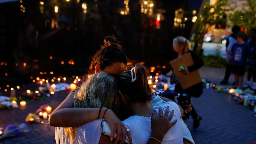 Three women hug, surrounded by candles on the ground, glowing in the evening light