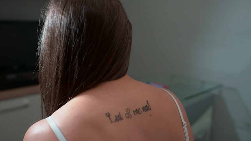 The back of a woman with straight brown hair, showing her upper back tattoo