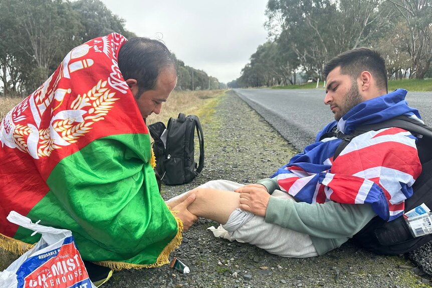 A man in Afghan flag rubs the leg of a man in an Australian flag by the side of the road.