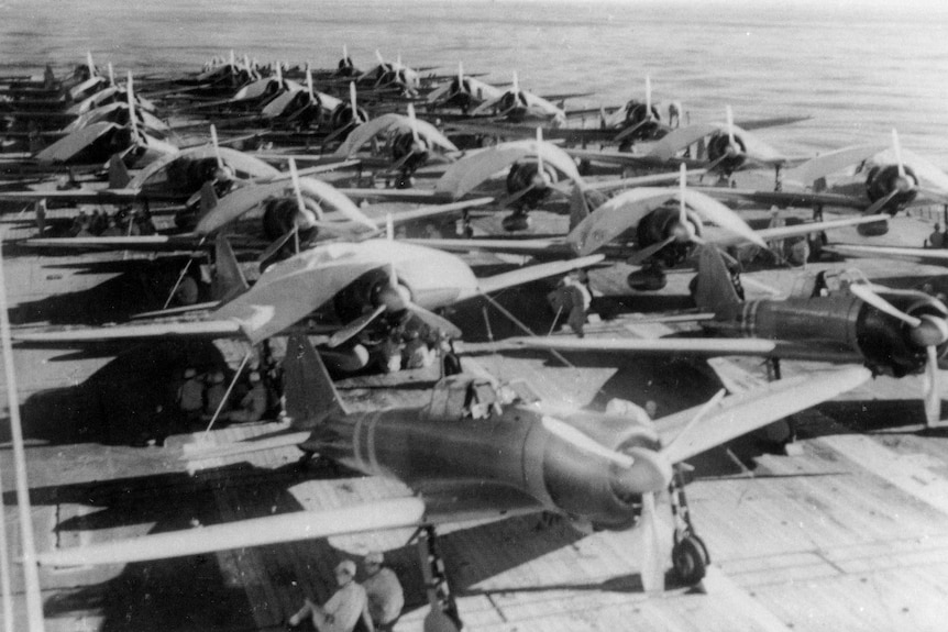 A black and white photo shows Japanese Imperial Air Force crewmen service aircraft on a carrier during World War Two