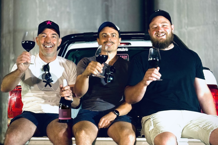 Three men sit on the back of a ute tray in a concrete warehouse raising wine glasses with red wine and smiling