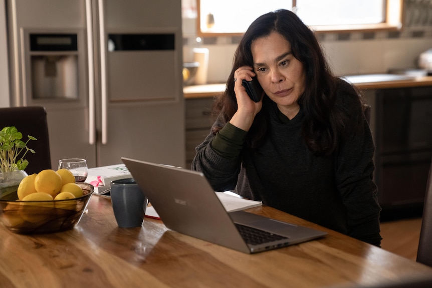 A woman sits in front of a laptop with a phone to her ear.