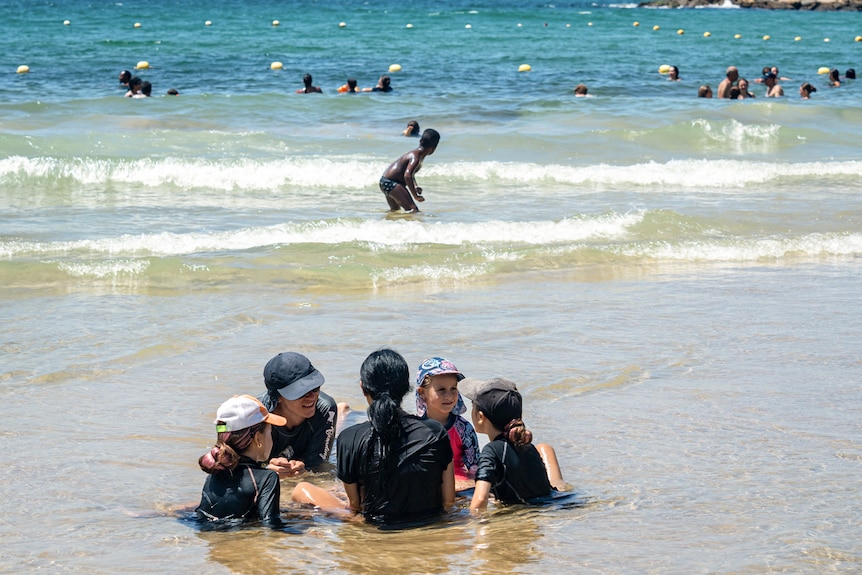 A group of kids sit in a circle in shallow water on the beach while other people swim further out in the ocean.