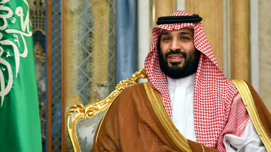 Saudi Arabia's Crown Prince Mohammed bin Salman sits next to a Saudi flag wearing a keffiyeh.