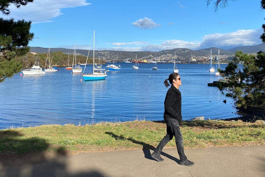 A man in jeans, black shirt and with a black ponytail walks in front of a bay where there are sailboats.