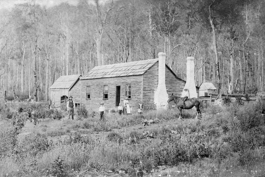a black and white photo of an old timber house in the bush, the family are all outside including someone on horseback