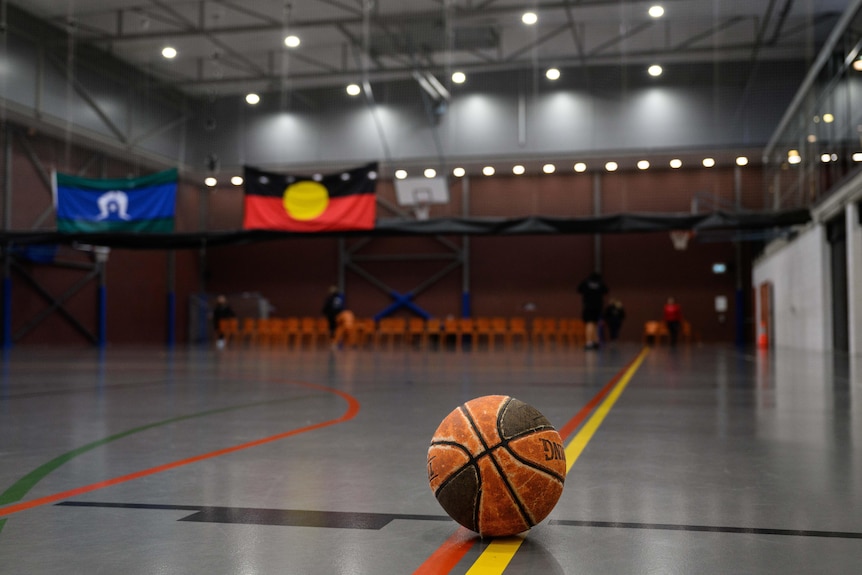 Баскетболна топка стои в празна зала с местно знаме, окачено на заден план