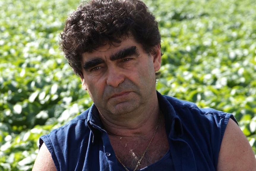 A close-up of Tony Galati in a blue shirt and wearing a gold chain, with fields in the background.