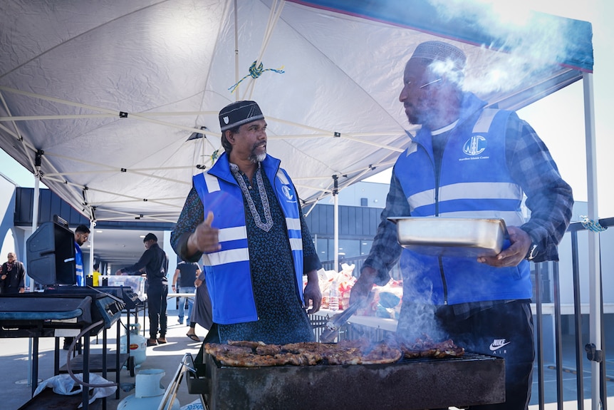 Two men in blue vests barbecuing meat
