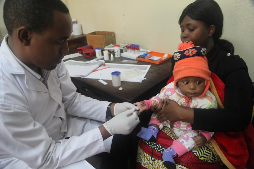A man in a white coat takes blood from an infant wearing an orange hat while being held by a woman. 