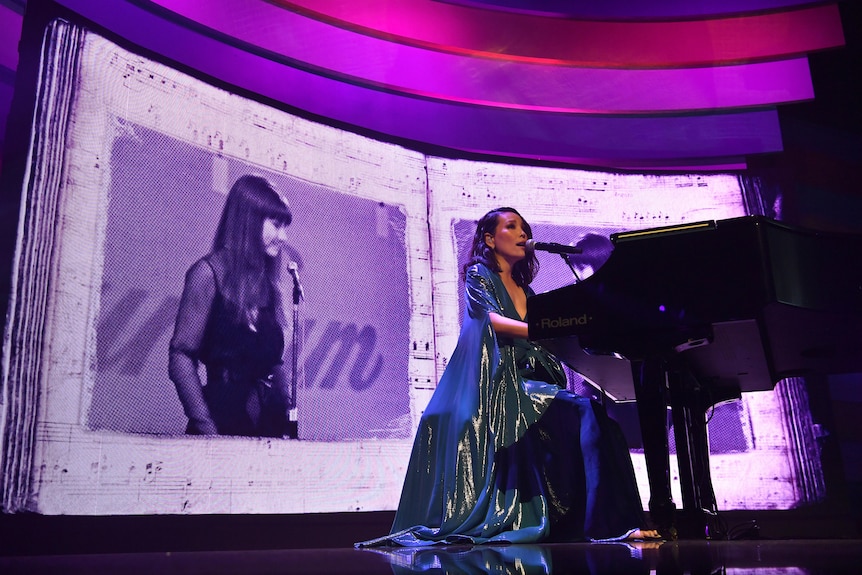 dami im sings into a microphone and plays piano in front of a large screen showing a picture of judith durham