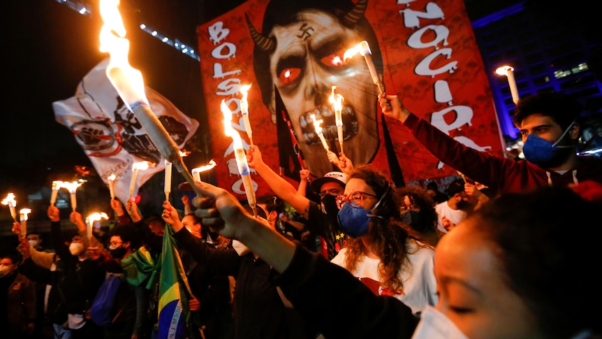 Protesters hold flames in front of a photo of Bolsonaro drawn as the devil