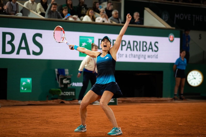 A women's tennis player screams and throws her arms wide in the air as she celebrates a victory at the French Open. 