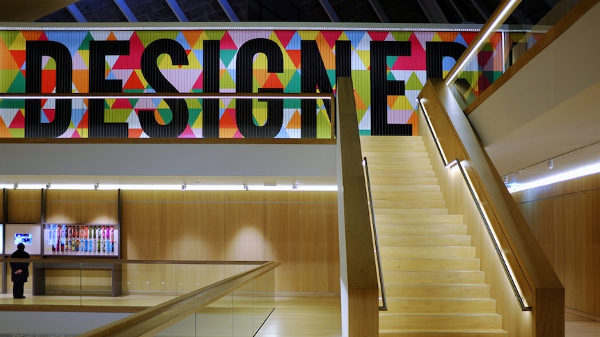 You view a warm wooden museum lobby with stairs rising up to a sign that reads, designer.