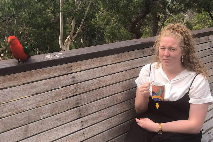 Tess Roberts sits on a bench with a mug in her hand looking unimpressed, with leafy trees behind her.