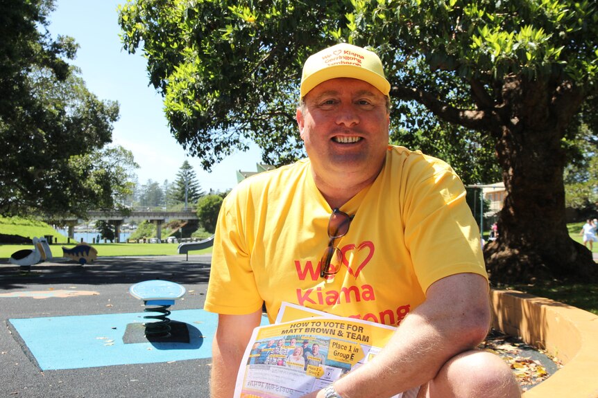 A smiling man sits near a playground in his yellow campaign hat and T-shirt, holding fliers.
