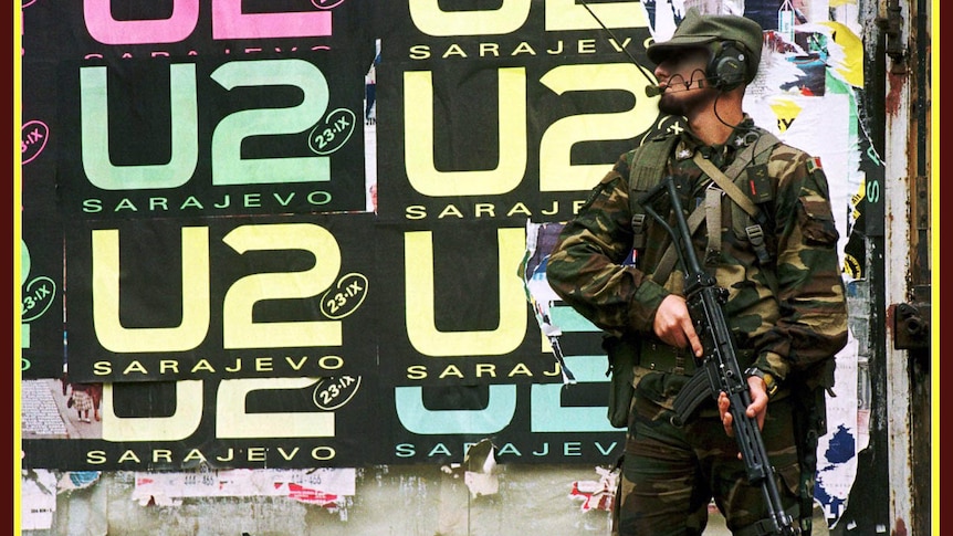 An armed Bosnian soldier stands watch by a wall plastered in concert posters for "U2 Sarajevo"