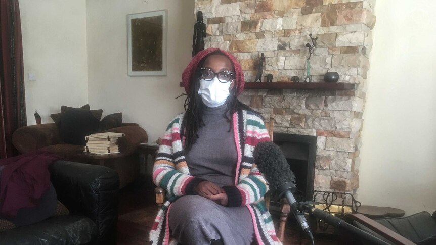 Zimbabwean novelist Tsitsi Dangarembga sits in her living room wearing a face mask and a beanie.