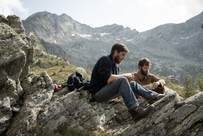 A fair-haired Italian man and a dark-haired Italian man sit on rocks in a mountainous landscape eating bread with their hands.