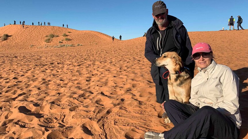 A dog sits on a sand dune between a man and a woman.