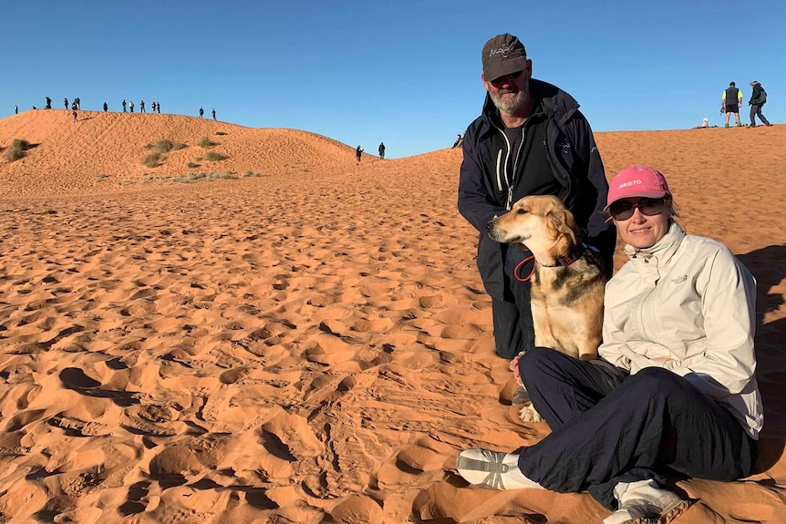 A dog sits on a sand dune between a man and a woman.