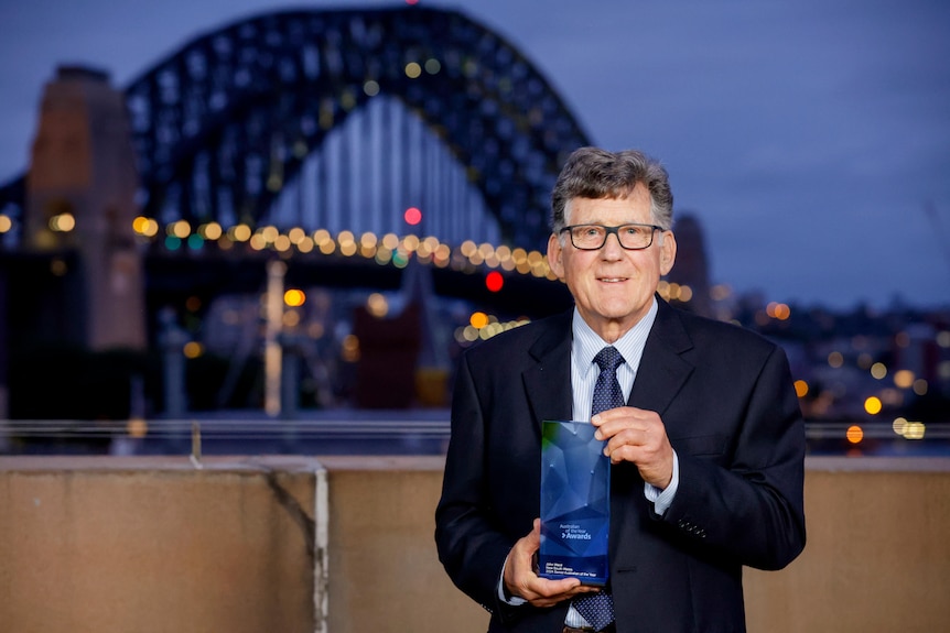 A man with grey hair and glasses holds up a glass award with the Sydney Harbor Bridge in the background.
