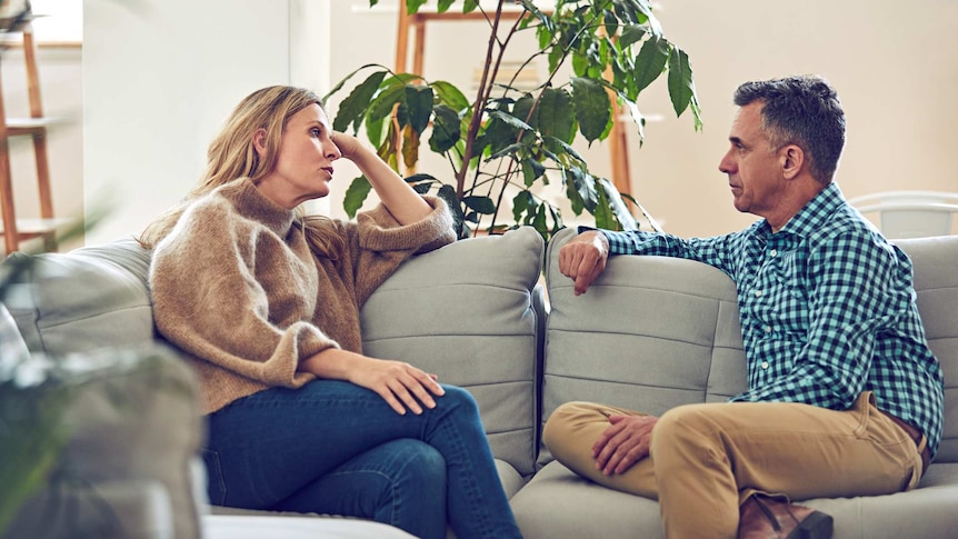 A middle age couple sitting on a grey couch and talking to each other.