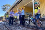 Three politicians stand on railway tracks in front of an old platform, holding a map and thumbs up, flanked by cyclists